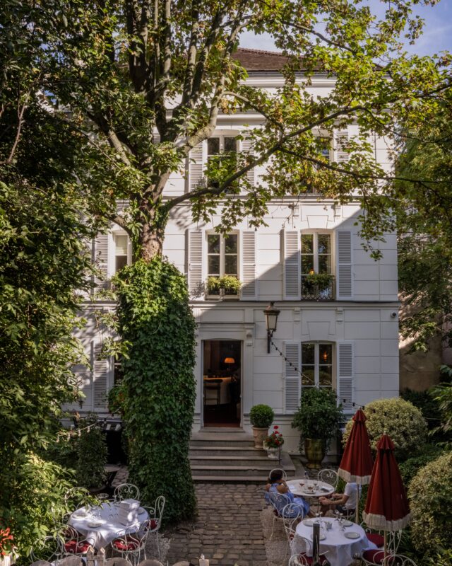 Elegant white facade of Hôtel Particulier Montmartre surrounded by greenery, with outdoor seating and red umbrellas.