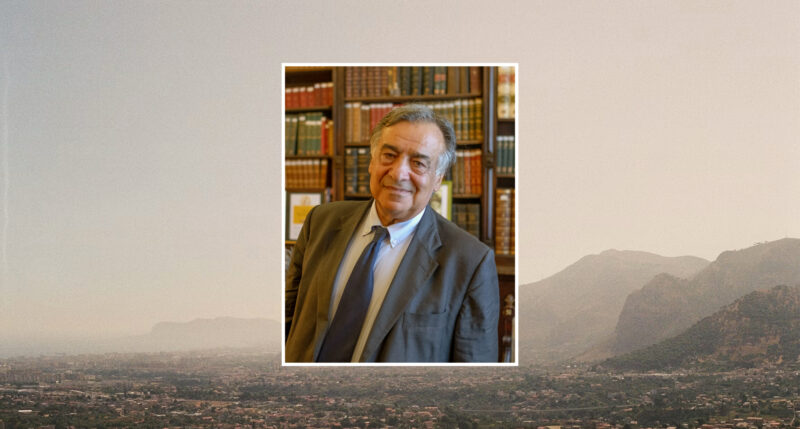 Portrait of a man in a suit against a blurred landscape of Palermo's mountains and cityscape.
