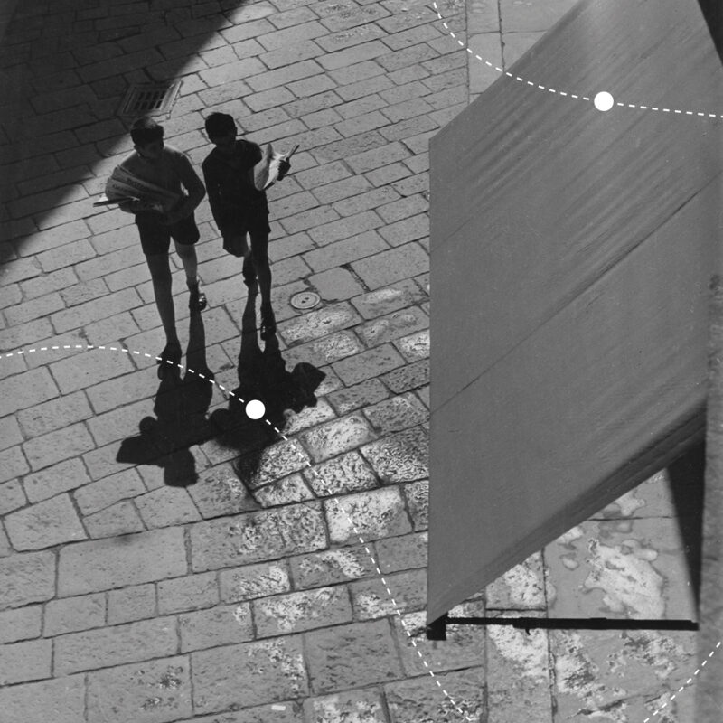 Two boys walking on a cobblestone street, casting long shadows, with a partially visible wall on the right.