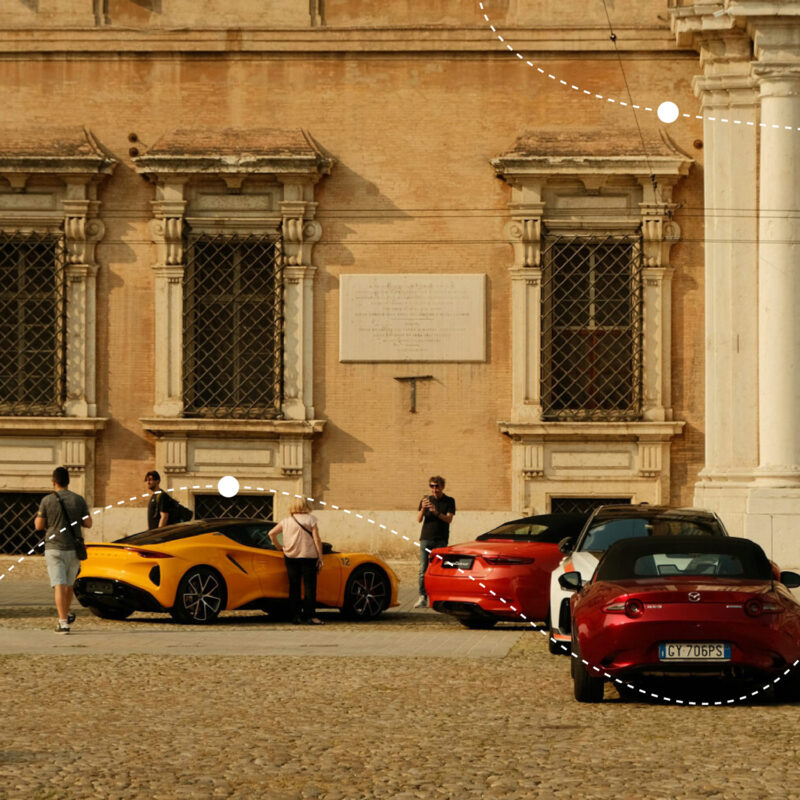 Four cars, including a yellow sports car and a red convertible, parked near a historic building with people gathered around.