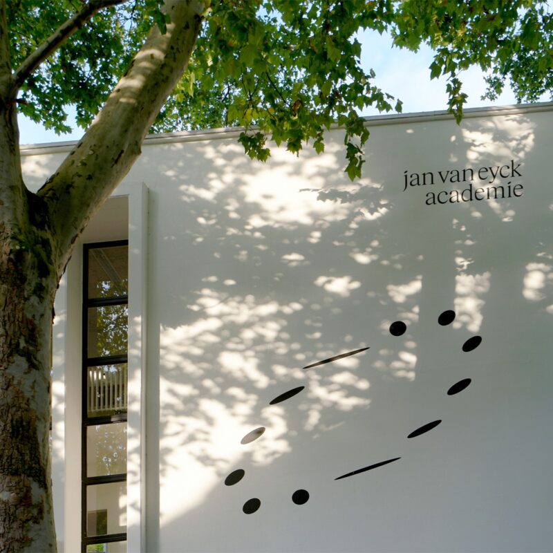 White wall of the Jan van Eyck Academie building with a modern black circular design and green tree branches above.