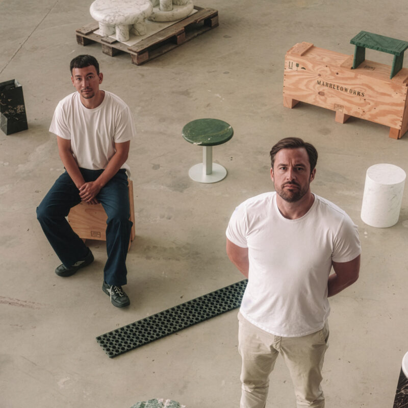 Two men in white t-shirts stand and sit on stools in a spacious workshop, surrounded by various sculptural objects.