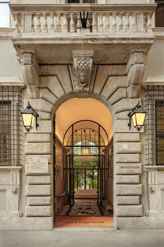 Elegant archway of Palazzo Talìa's main entrance, featuring decorative stonework and lanterns, leading to a well-lit inte...