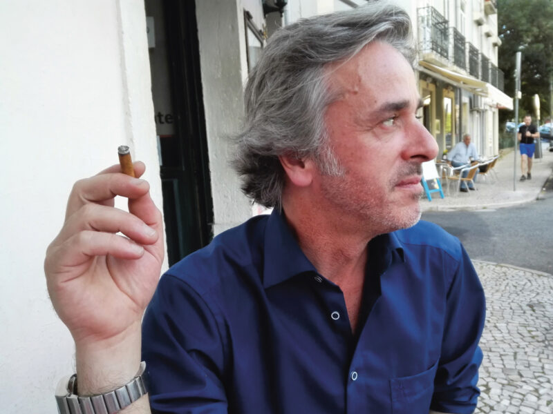 Man with gray hair and a blue shirt holding a cigarette, gazing thoughtfully to the side in a Lisbon street.