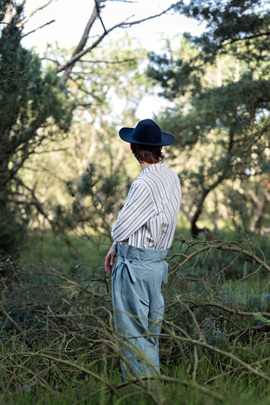 Person in a striped shirt and wide-brimmed hat stands among trees and underbrush, facing away from the camera.