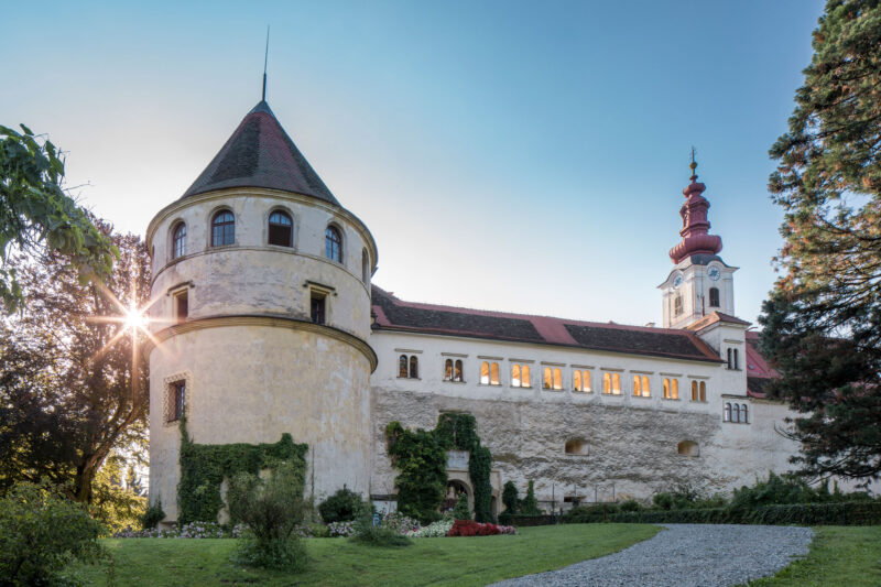 Historic Schloss Hollenegg with a tower, illuminated windows, and a sunset glow in the background.
