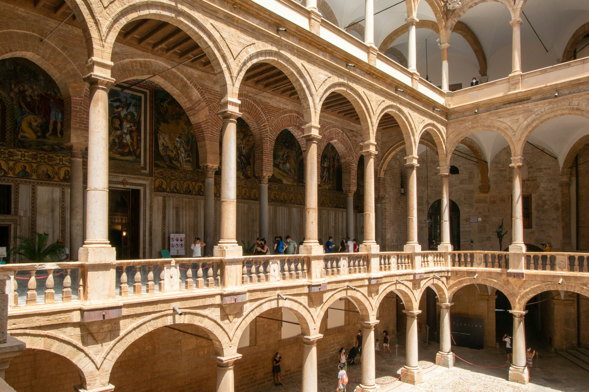 Historic courtyard with arched columns and a balcony, filled with visitors, showcasing Palermo's architectural beauty.
