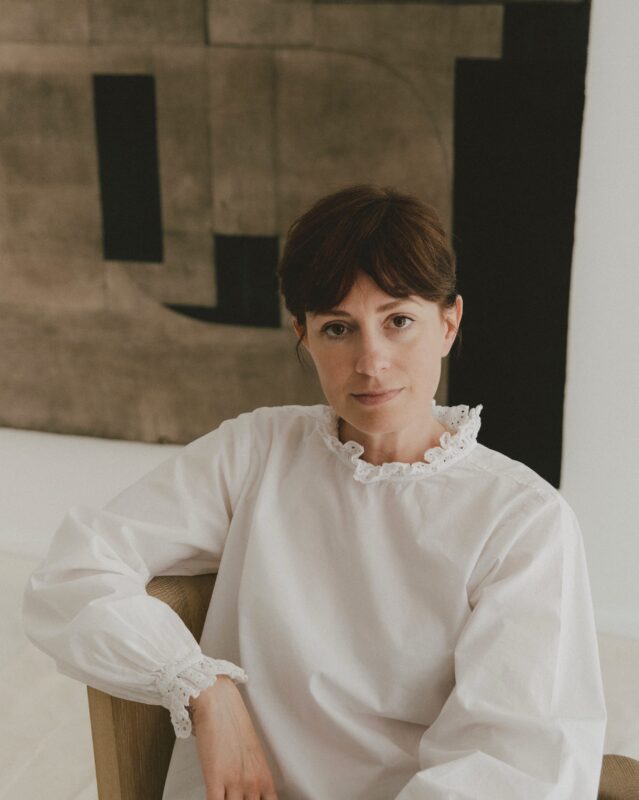 Woman in a white blouse with ruffled cuffs sits in front of an abstract artwork, gazing directly at the camera.