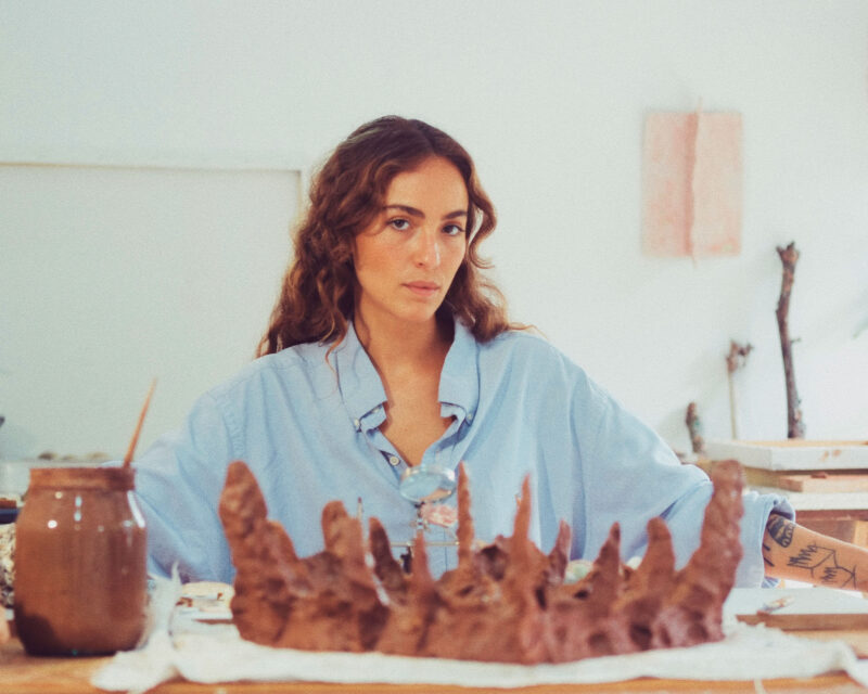 Young woman with curly hair, wearing a blue shirt, sitting at a table with clay sculptures and art supplies.