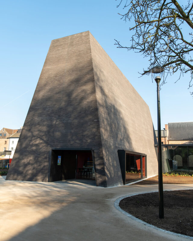 Modern angular building with a textured dark facade, surrounded by a landscaped area and a street lamp.