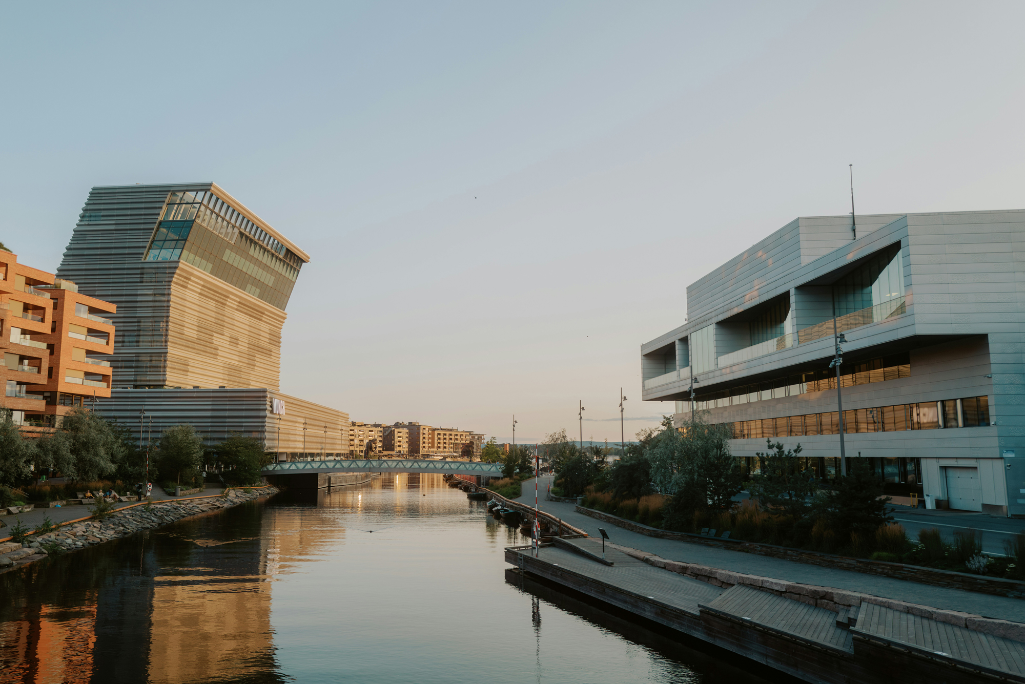 Modern buildings reflect in a calm river at sunset, with a pedestrian walkway lined by trees and people in the distance.