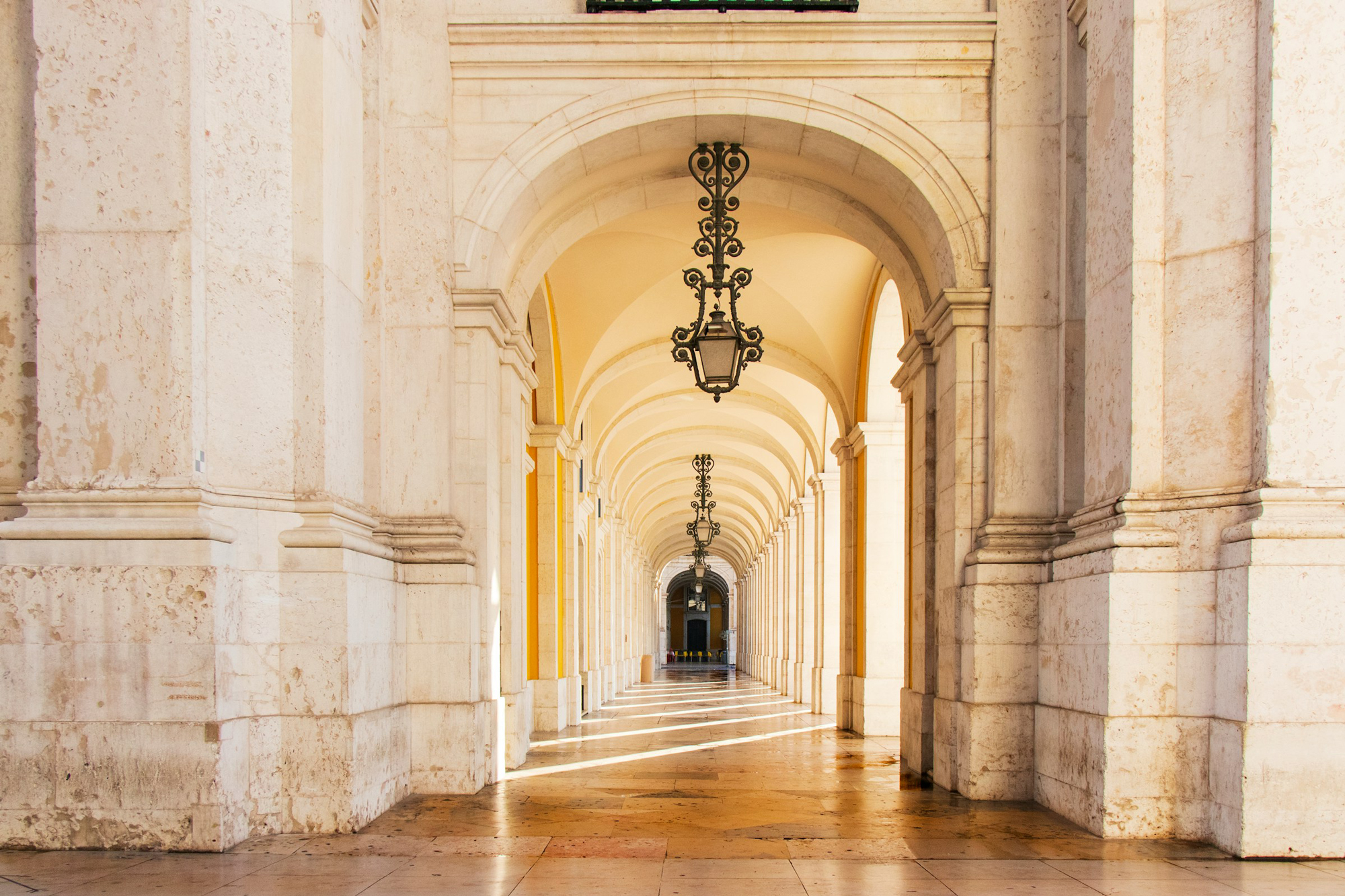 Arched corridor with ornate lanterns, illuminated by sunlight, showcasing intricate stonework in Lisbon.
