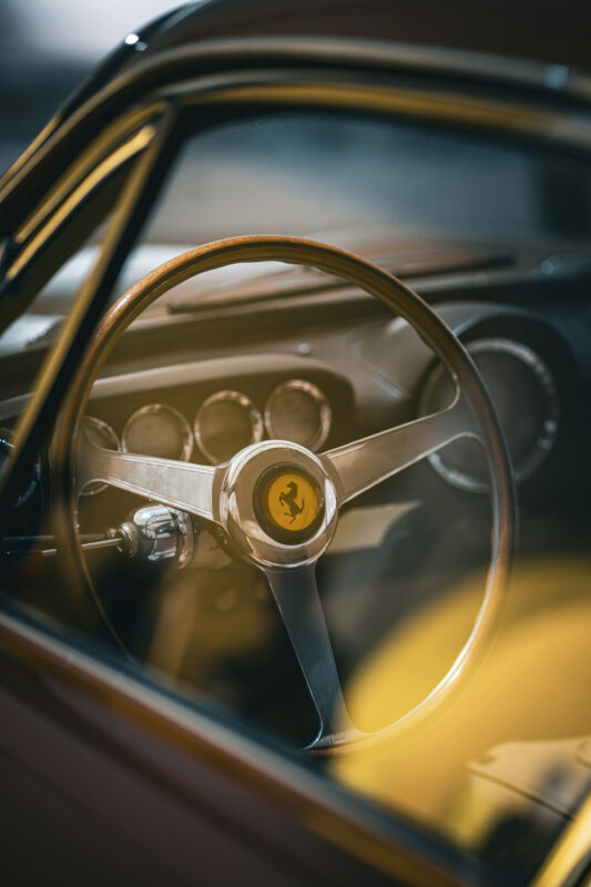 Ferrari steering wheel with a wooden rim, showcasing the iconic logo and dashboard dials in soft focus.