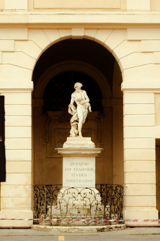 Statue of a man holding a child, set in an archway with a decorative railing and Latin inscription on the pedestal.