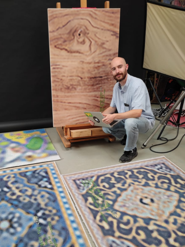 Portrait of Jonas Hejduk sitting on the floor next to patterned rugs, with a wooden panel on an easel behind him.