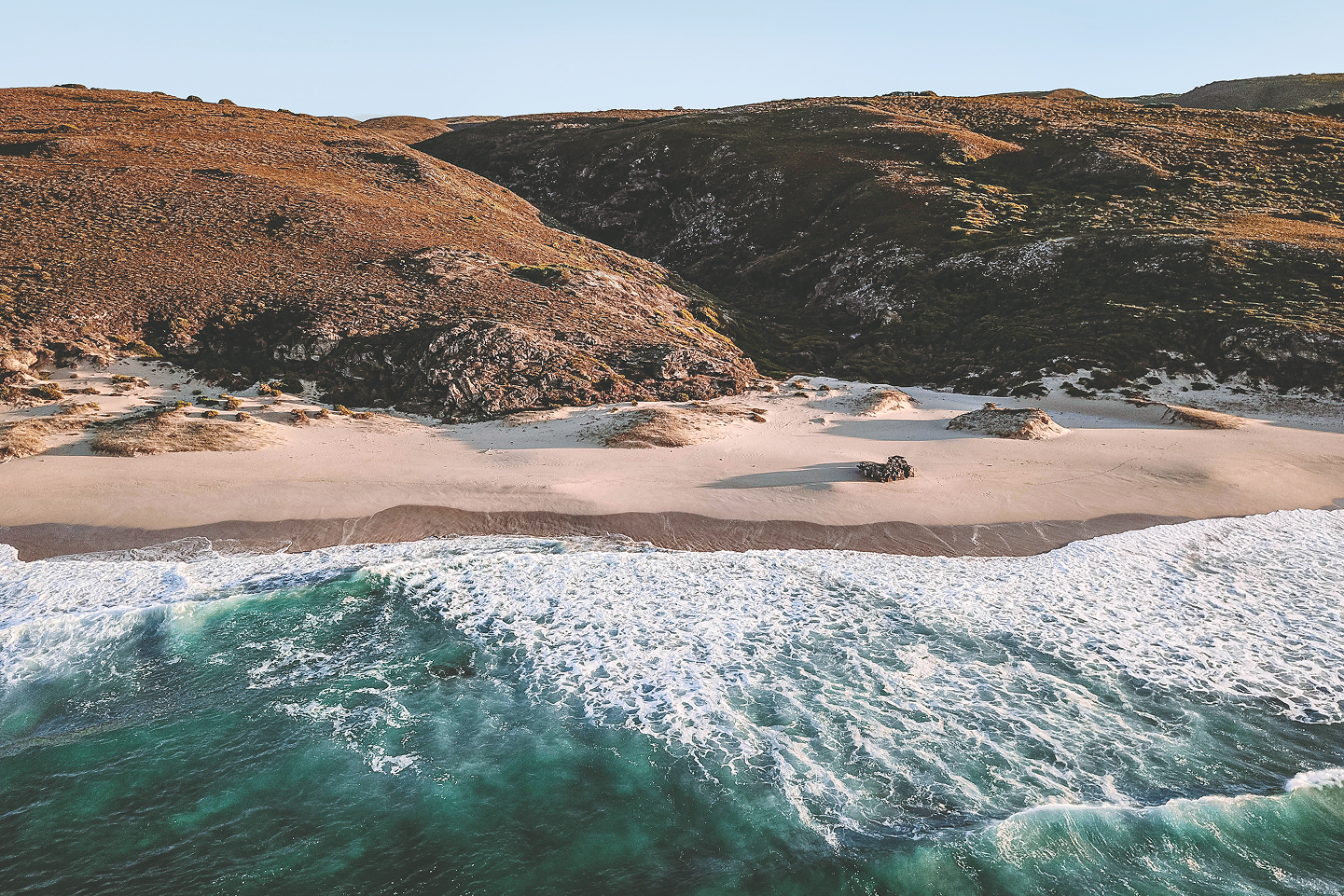 Aerial view of a sandy beach with gentle waves, surrounded by rocky hills and greenery.