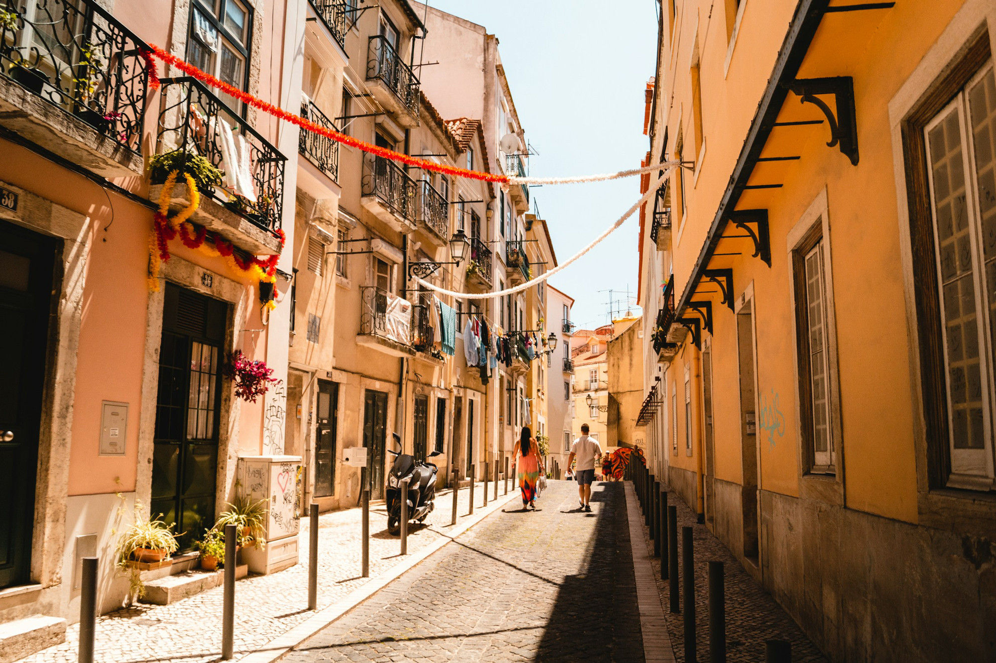 Narrow cobblestone street in Lisbon, lined with colorful buildings and hanging laundry, with two people walking.