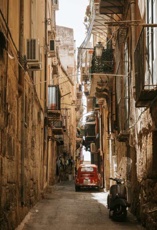 Narrow cobblestone street in Palermo, lined with old buildings, featuring a red car and a scooter parked nearby.