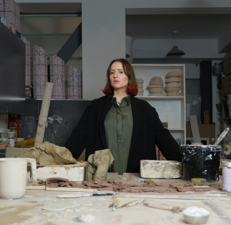 Woman with shoulder-length hair, red tips, stands confidently in a pottery studio surrounded by clay and tools.