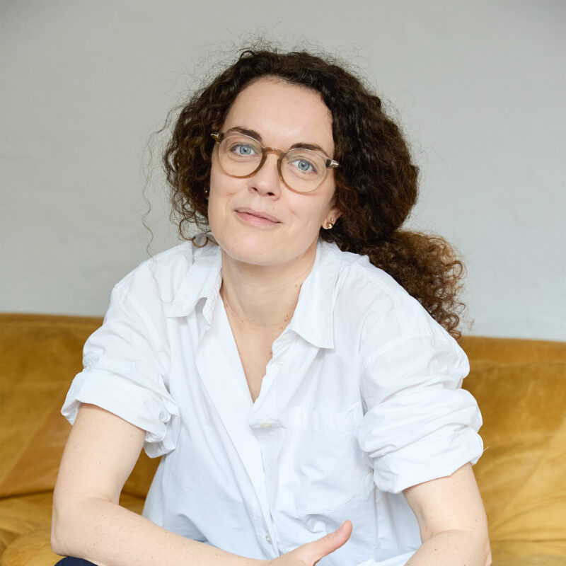 Woman with curly hair and glasses, wearing a white shirt, sitting on a yellow couch, smiling softly.