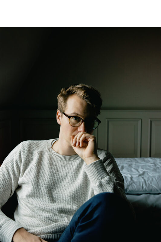 Young man in a light sweater and glasses, resting his chin on his hand, seated in a dimly lit room.