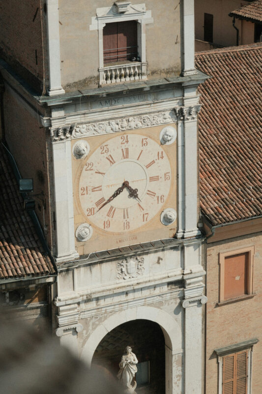 Clock tower with a large clock face, featuring Roman numerals, set against a backdrop of terracotta rooftops.