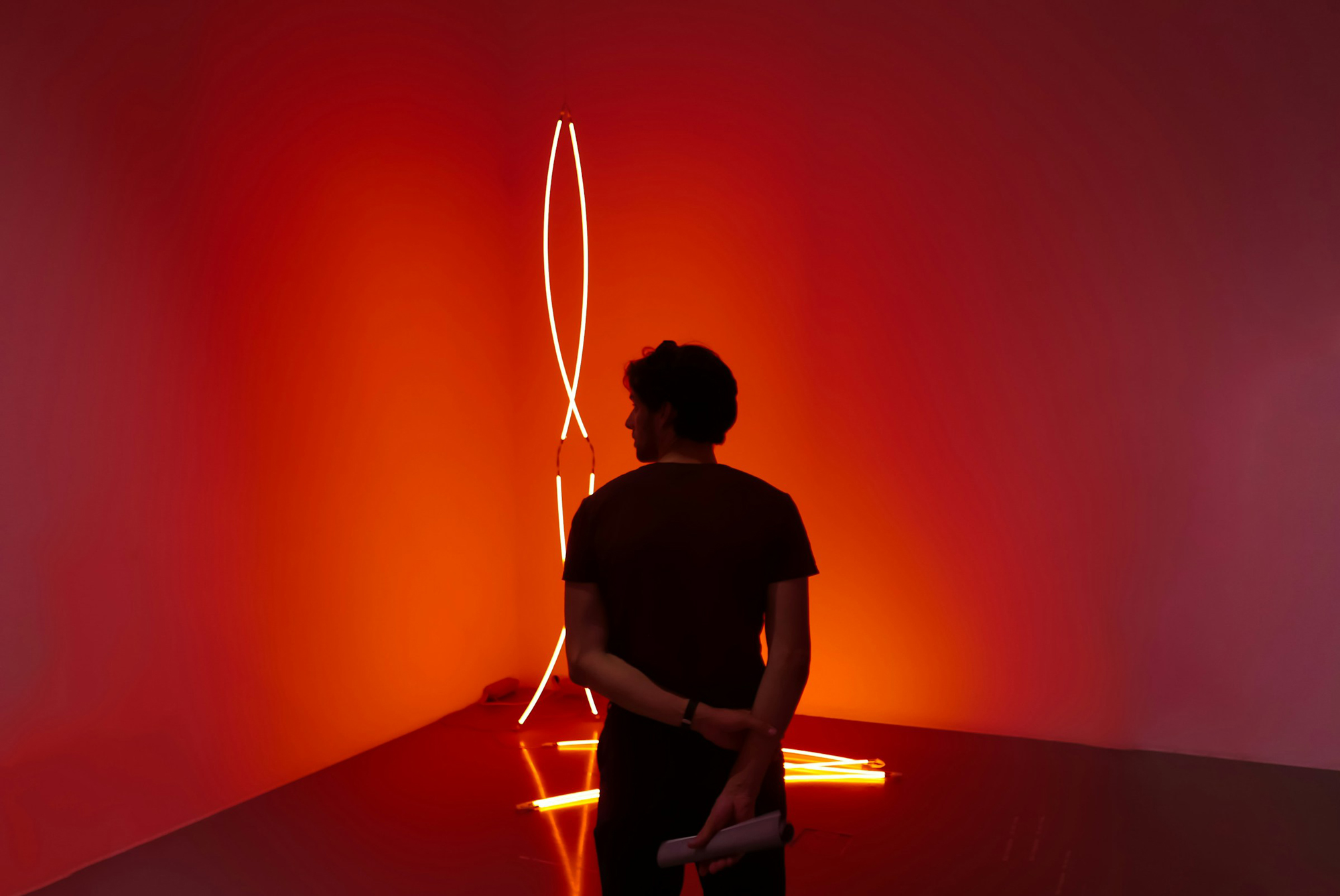 Person standing with back to the camera, observing glowing neon light installation against an orange background.