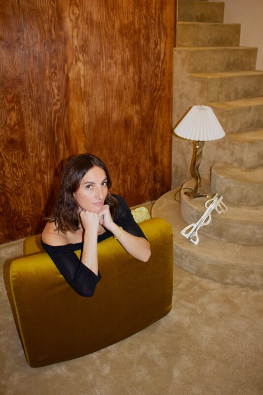 Woman with long brown hair resting her chin on her hands, seated on a green velvet couch near a staircase and lamp.