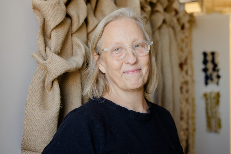 Portrait of Annie Coggan smiling, wearing glasses and a black shirt, with textured fabric in the background.