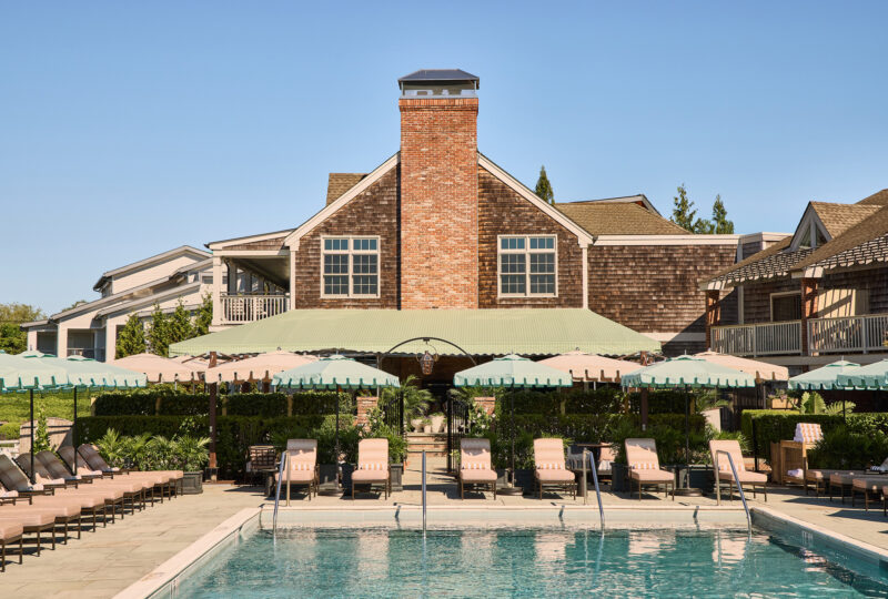 Pool area with lounge chairs and green umbrellas, featuring a brick building in the background under a clear blue sky.