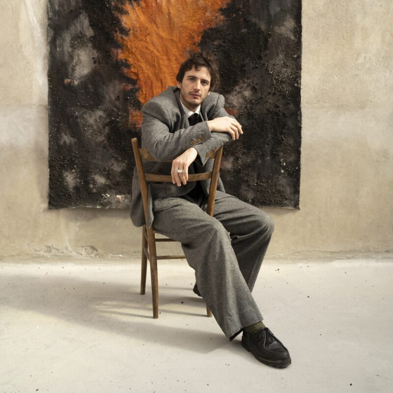 Man in a gray suit sitting on a wooden chair against an abstract artwork with orange and black tones.