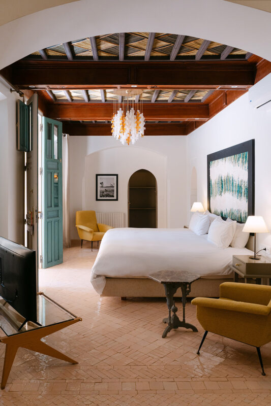 Bright hotel room featuring a king-sized bed, yellow armchairs, a TV, and a decorative chandelier, with open doors reveali...