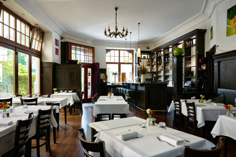 Elegant restaurant interior with white tablecloths, wooden chairs, and a bar, featuring large windows and floral centerpie...