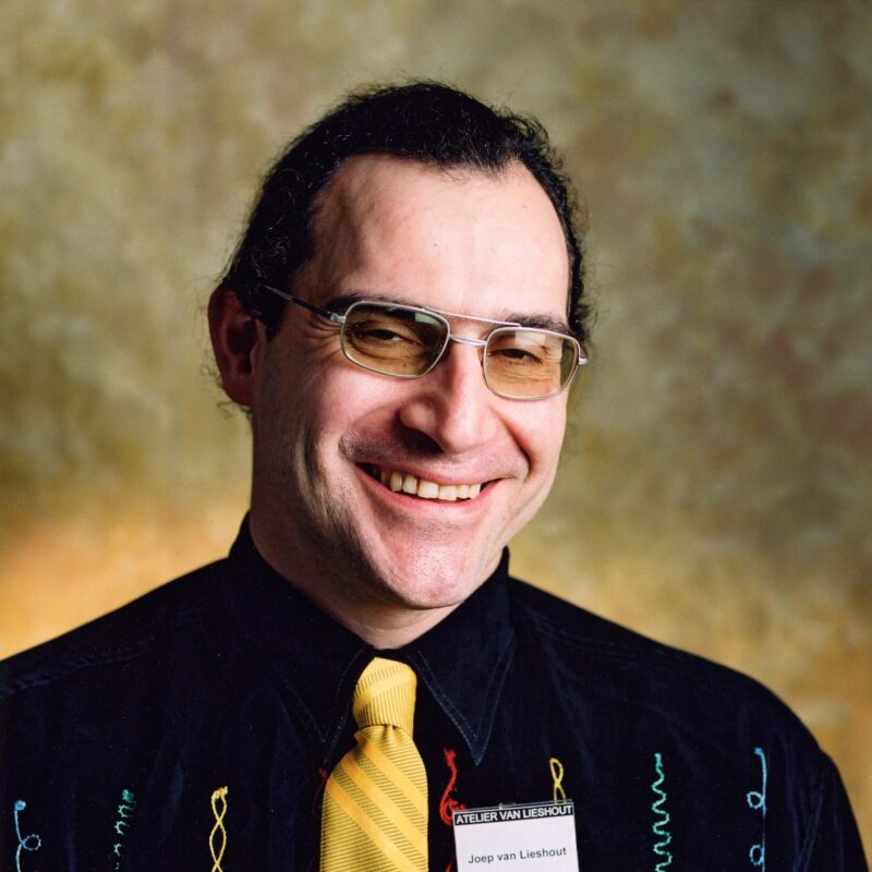 Smiling man with glasses and a yellow tie, wearing a black shirt with colorful embroidery, against a blurred background.