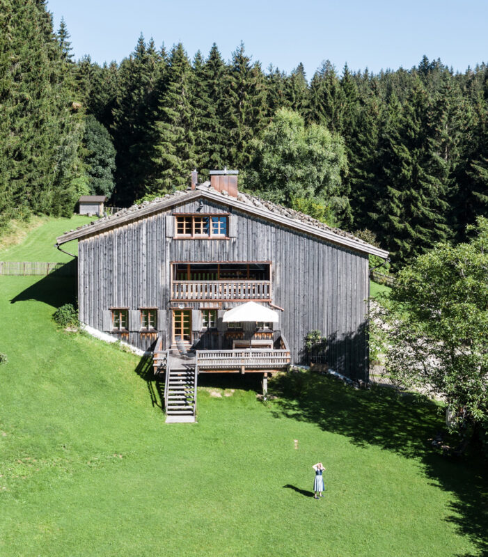Wooden house surrounded by green grass and trees, with a person standing in front, under a clear blue sky.