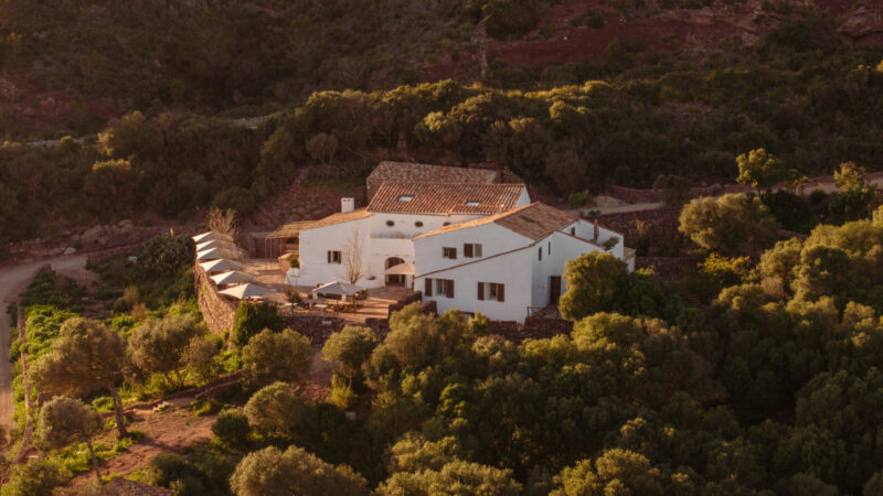 Aerial view of a white house surrounded by greenery and rocky terrain, with a pathway leading to it.