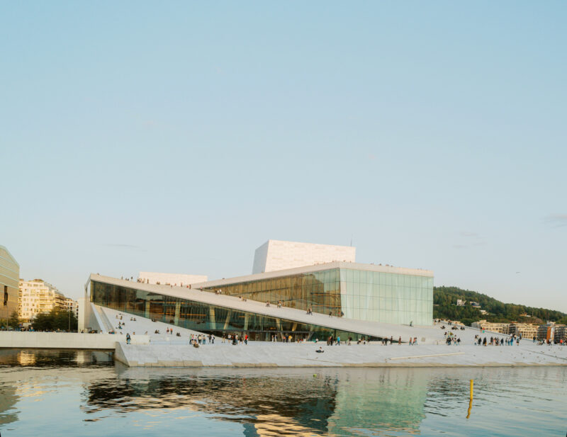 Modern architectural structure with a sloping roof, situated by water, reflecting the Oslo skyline at sunset.