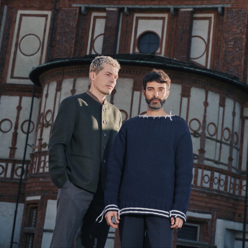 Two models stand in front of a historic building, wearing stylish knitwear in dark tones, showcasing contemporary fashion.