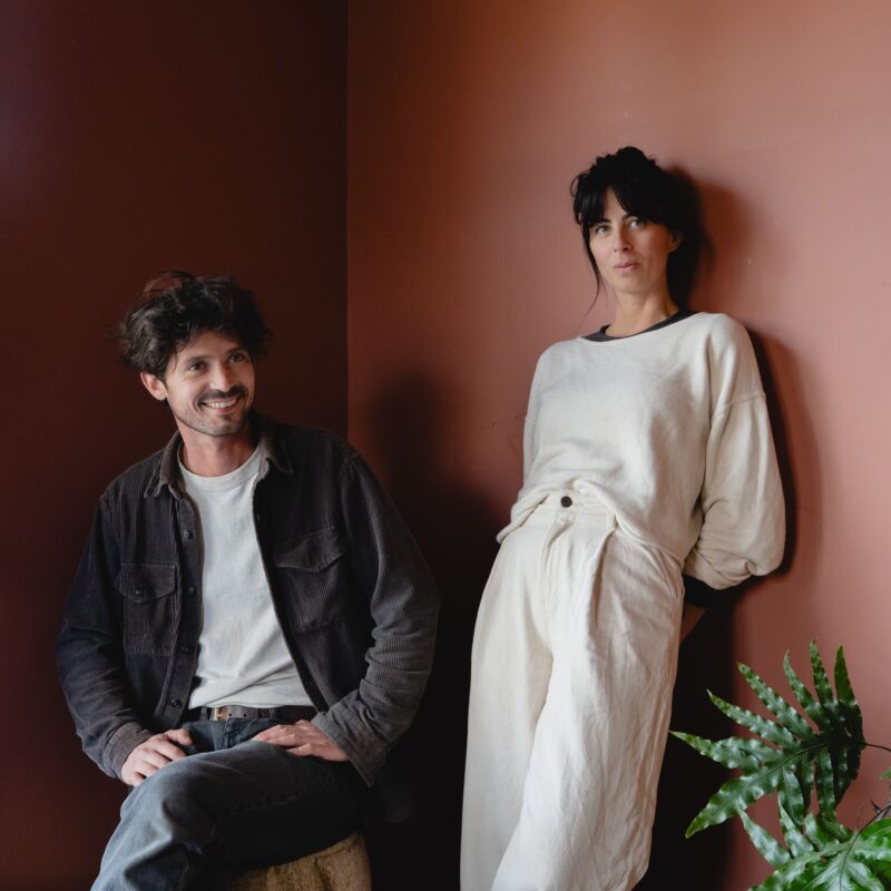 A man and a woman pose together in a cozy room with a warm, earthy-toned wall and a potted plant.