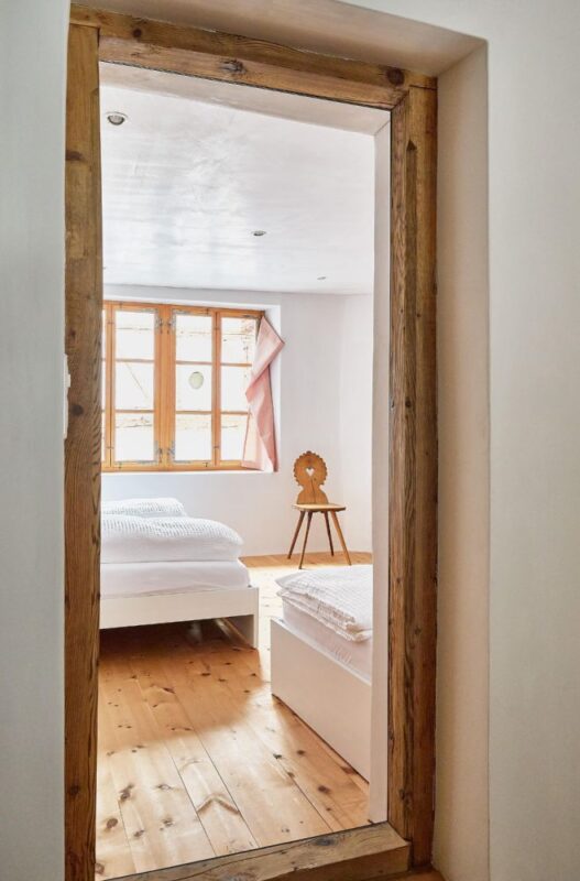 Bright interior view through a wooden door frame, showcasing a cozy bedroom with two beds and a wooden chair.