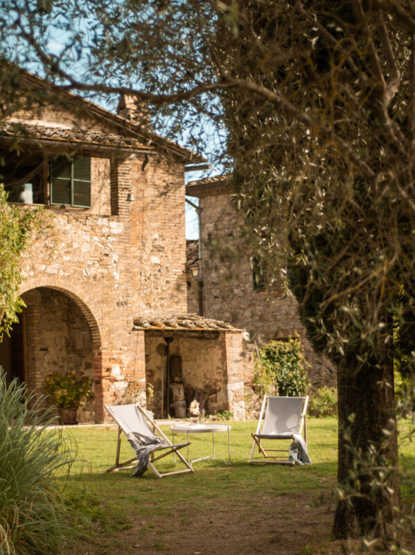 Two white folding chairs and a small table in a grassy area, framed by olive trees and rustic stone buildings.