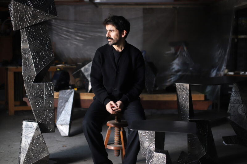 Portrait of André Teoman seated on a stool in a workshop, surrounded by sculptural metal pieces.