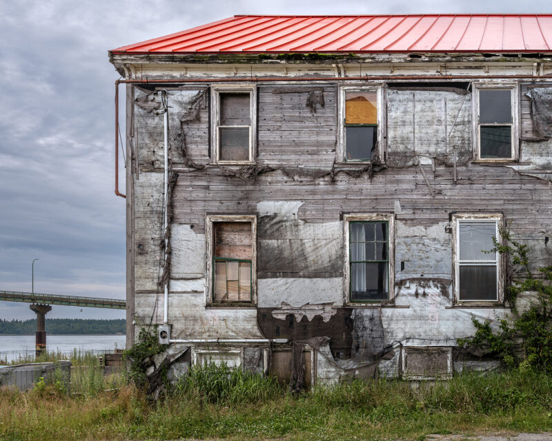 Weathered wooden house with a red roof, showing peeling paint and boarded windows, near water in Lubec, Maine.