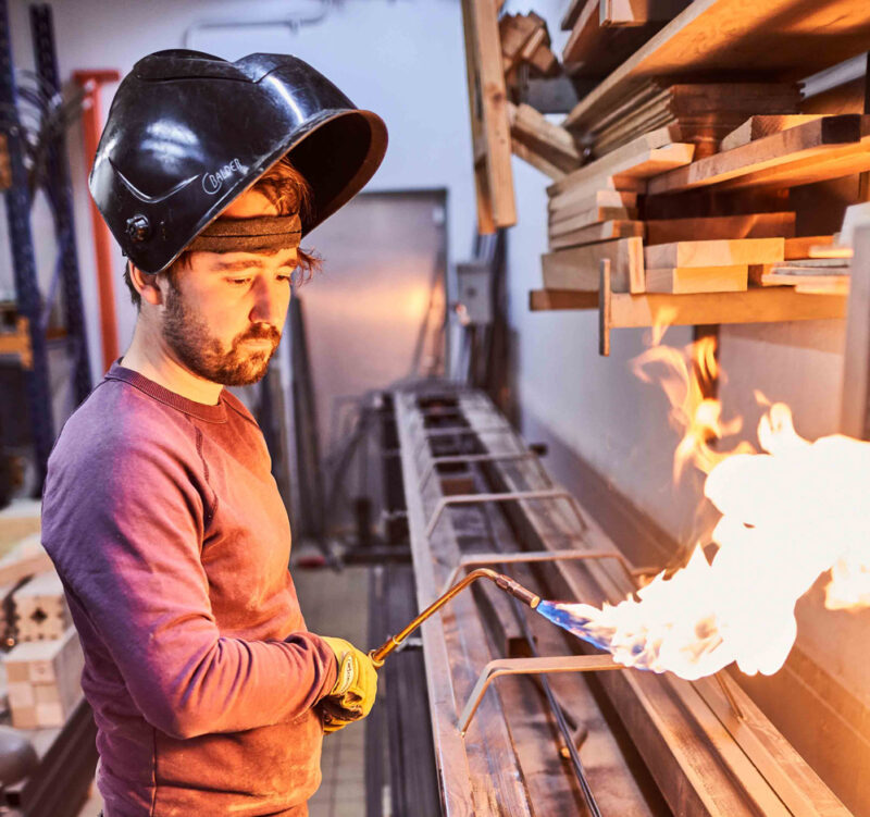 Man in a welding helmet uses a torch to heat metal in a workshop, surrounded by wooden materials.