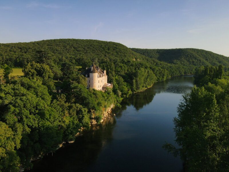 Château de la Treyne overlooking a river, surrounded by lush greenery and hills under a clear sky.